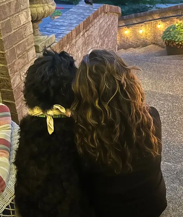 A person with long curly hair and a black curly-coated dog sitting side by side, seen from behind on brick steps